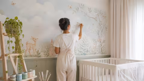 Artista pinta mural suave com animais e nuvens em quarto de bebê.